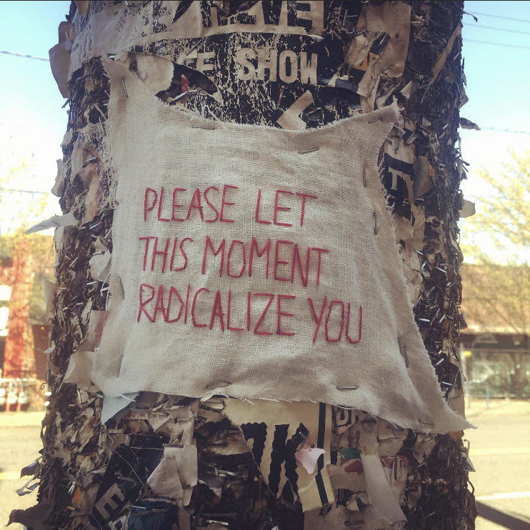A white asymmetrical piece of fabric is stapled to a black utility pole and stitched on to it in red thread is the Alexandria Ocasio-Cortez quote "Please let this moment radicalize you."
