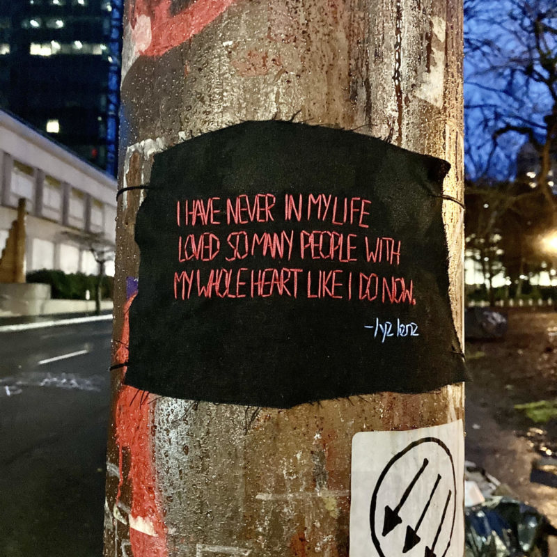 A black rectangular piece of fabric is stapled to a wooden utility pole and stitched on to it in red thread is a quote from Lyz Lenz's 2018 essay on The Rumpus titled "Why Writing Matters in the Age of Despair." The quote reads "I have never in my life loved so many people with my whole heart like I do now."